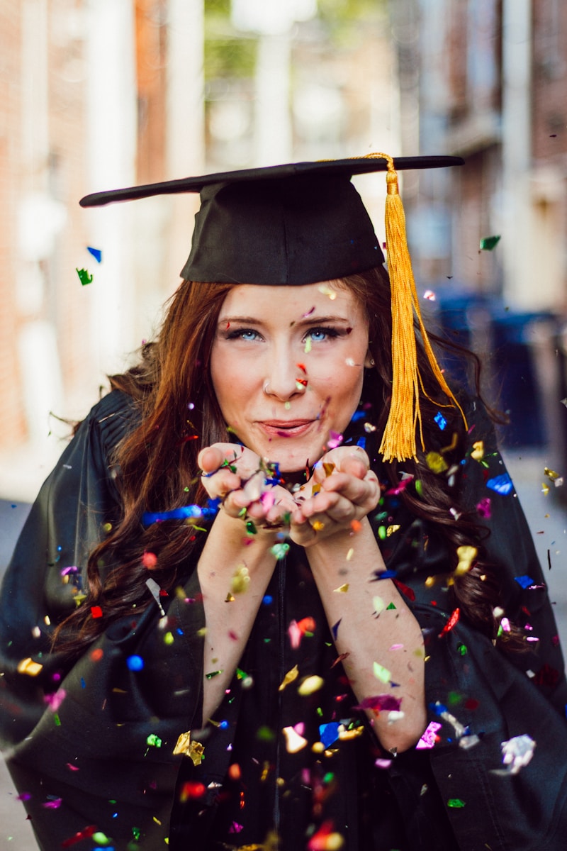 A joyful nursing graduate from one of the best nursing schools in Virginia, in a cap and gown, blows colorful confetti towards the camera on a sunny day.